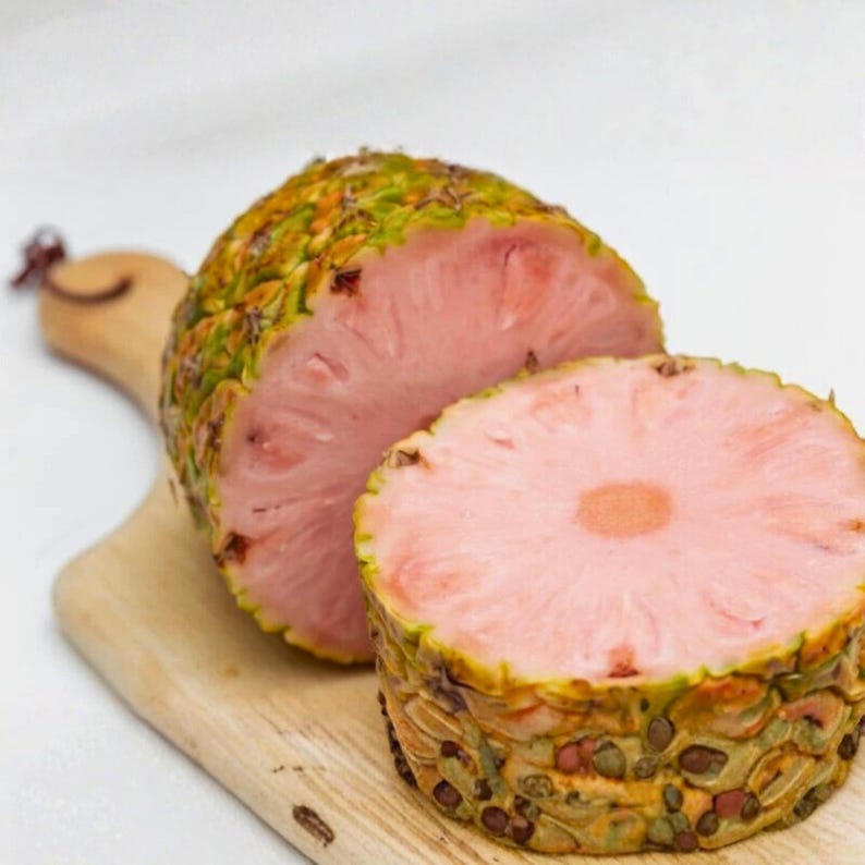 Puede incluir: Una pi&ntilde;a rosa cortada por la mitad con un exterior texturizado verde y marr&oacute;n, sobre una tabla de cortar de madera. El corte revela la vibrante pulpa rosa y un centro.