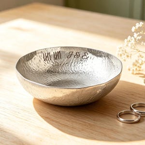 May include: A round, silver-toned metal bowl with a textured, hammered surface. The interior rim is engraved with the text "III III J & E". Two plain, thin silver-toned metal rings are positioned near the base of the bowl.
