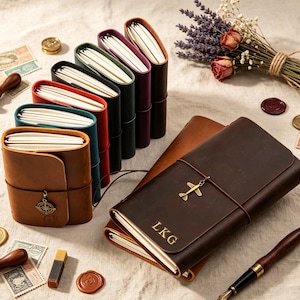 May include: A collection of leather journals in various colors including brown, teal, red, black, and purple. One dark brown journal features the letters LKG and an airplane charm. Another brown journal has a compass charm and the letters GR.