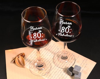 May include: Two clear wine glasses filled with red liquid, each with the text "Susan 80 & Fabulous" in white script. The glasses sit on a piece of paper with text, accompanied by decorative items and gray stone cubes.