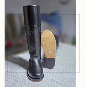 May include: A tall black leather boot with a smooth finish and a rounded toe. Beside it stands a second boot, showing the tan-colored sole and a black stacked heel. The boots feature a clean, structured design with visible vertical stitching.