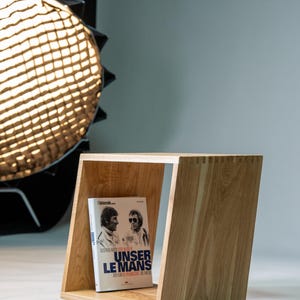 Könnte beinhalten: Ein helles Holzregal mit schräger Konstruktion. Es zeigt ein Buch mit dem Titel "UNSER LE MANS" mit zwei Männern auf dem Cover. Das Regal ist aus hellem Holz gefertigt und weist sichtbare Holzverbindungen auf.