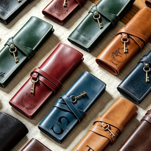 May include: A collection of rectangular leather pencil cases in various colors including black, red, green, blue, and tan. Each case features a wrap-around leather strap secured with a metal key charm and personalized engravings like names, dates, and floral or animal motifs.