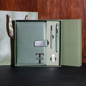 Könnte beinhalten: Ein salbeigrünes Geschenkset mit einem Notizbuch, einem Stift und Zubehör, präsentiert in einer passenden Box. Das Notizbuch ist mit dem Buchstaben "T" und dem Namen "Theodore" geprägt. Eine Geschenktüte ist enthalten.