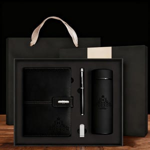 Könnte beinhalten: Ein schwarzes Geschenkset mit Notizbuch, Stift, Becher und Büroklammer, alles in einer schwarzen Box angeordnet. Das Notizbuch hat einen Verschluss und das Wort "HUDSON" ist auf den Artikeln aufgedruckt. Eine schwarze Geschenktüte mit weißem Griff befindet sich hinter der Box.