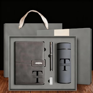 May include: A grey gift set containing a notebook, pen, flask, and USB drive, all personalised with the name "Theodore." Presented in a grey box with a matching gift bag, ideal for a special occasion.