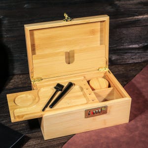 May include: A light brown bamboo box with a hinged lid, open to reveal compartments and accessories. The box includes a tray, a small brush, a black tube, and a wooden container. A combination lock is visible on the front.