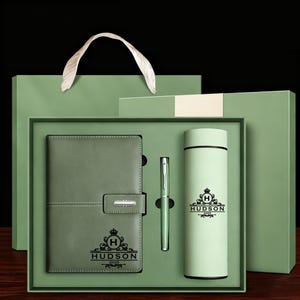 May include: A gift set in a green box, featuring a notebook, pen, and insulated tumbler. The notebook has a clasp and the word "HUDSON" printed on it. The tumbler and notebook have a matching logo. The set is presented in a green gift bag with a white ribbon handle.