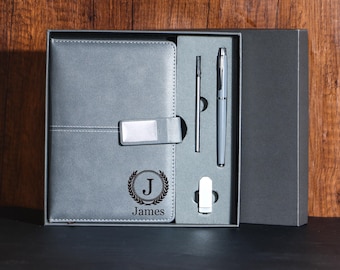 May include: A gray leatherette journal and pen set in a gift box. The journal has a silver clasp and the letter "J" with the name "James" in a laurel wreath. The set includes a pen, a stylus, and a pen.