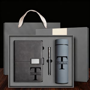 May include: A grey gift set containing a notebook, pen, and a tumbler, each bearing the initial "T" and the name "Theodore". The set is presented in a grey box with a matching gift bag, suitable for a personalised gift.