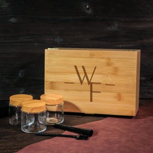 Puede incluir: Una caja de bambú con las letras "WF" grabadas y tres frascos de vidrio con tapas de madera. Una herramienta de metal negro y un tubo negro están frente a los frascos. El fondo es de madera oscura.