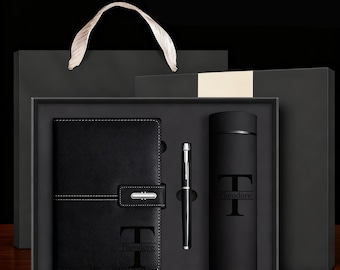 May include: A black gift set containing a leather-bound notebook, a pen, and a thermos. The notebook and thermos feature the name "Theodore" and a large "T" initial. The set is presented in a black box with a gift bag.