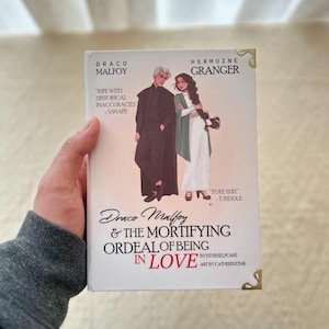 Könnte beinhalten: Buchcover mit Draco Malfoy und Hermine Granger. Der Titel lautet "Draco Malfoy & The Mortifying Ordeal of Being in Love". Die Illustration zeigt die Charaktere in einer romantischen Pose. Das Buch hat goldene Eckakzente.
