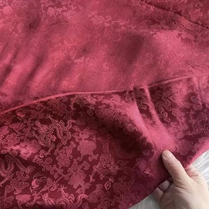 May include: Close-up of a burgundy fabric with a floral and dragon pattern. The fabric has a satin sheen and a detailed embossed design. The lower portion of the fabric features a more intricate dragon and floral motif.