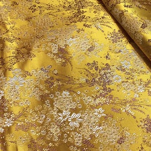 Puede incluir: Primer plano de una tela amarillo dorado con un estampado floral. El diseño presenta flores y follaje blancos y marrones, creando una apariencia texturizada y elegante. La tela está doblada, mostrando su caída y brillo.