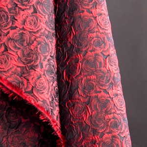 May include: Close-up of a fabric with a repeating rose pattern. The roses are a vibrant red colour, set against a dark background. The fabric has a textured appearance, with the roses appearing to be raised. The edge of the fabric has a red fringe.