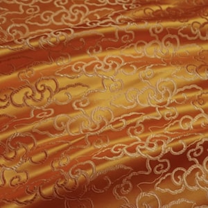 May include: Close-up of a golden-orange fabric with a swirling cloud-like pattern. The fabric has a satin sheen, with the design in a lighter, contrasting gold. The texture suggests a luxurious material, possibly for drapery or upholstery.