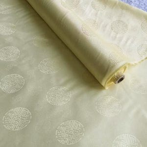 May include: A roll of pale yellow fabric with a repeating pattern of circular, stylized designs. The fabric has a subtle sheen and a soft texture. The design is a light cream color.