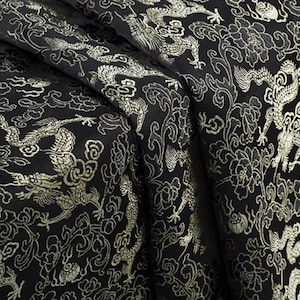 Op de afbeelding: Zwarte stof met een gouden draken- en bloemenpatroon. De stof is gedrapeerd en toont de ingewikkelde details van het ontwerp. De draken- en bloemenelementen zijn in de stof geweven.