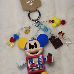 May include: A keychain featuring a cartoon mouse figure with blue ears and a red vending machine torso. The keychain includes charms of a chocolate bar, a yellow smiley face, a red heart, a blue heart, a yellow bow, and a yellow mouse head.