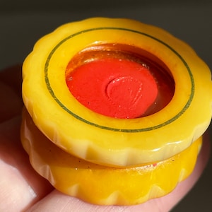 May include: A round, yellow, scalloped-edge button with a smooth, glossy finish. The center features a recessed, circular, bright red inlay surrounded by a thin, dark green circular line detail. The button has a thick, layered, disc-like profile.