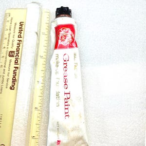 Può includere: Un tubetto di fondotinta Max Factor Grease Paint bianco con tappo nero, posizionato accanto a un righello giallo che mostra una lunghezza di circa 15 cm. Il tubetto presenta il marchio in rosso e oro e un peso netto di 56 grammi.