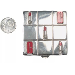 May include: A square silver-toned metal compact case featuring a tic-tac-toe grid design with red lipstick icons and red letter C accents. A United States dime is positioned to the left for scale, showing the compact is approximately 1.75 inches wide.