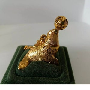 May include: A gold-toned metal pendant shaped like a seal balancing a ball on its nose. The seal has a textured surface finish and is attached to a small gold-toned chain link at the top. The pendant is depicted in a side profile view.