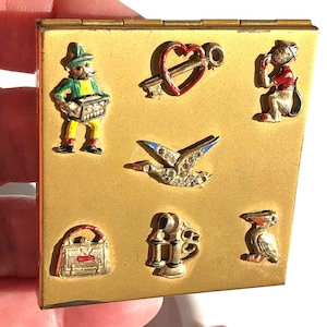 May include: A square gold-tone metal compact case featuring seven small decorative charms attached to the front surface. The charms include a person playing an accordion, a red heart with a key, a monkey, a bird with rhinestones, a handbag, a telephone, and a pelican.