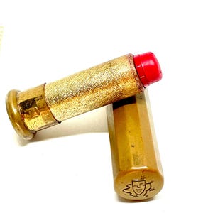 Può includere: Un rossetto vintage con un astuccio in metallo dorato testurizzato e una punta di colore rosso brillante. Il tappo esagonale dorato abbinato presenta un emblema inciso di una maschera teatrale circondata da piccole stelle sull'estremità.