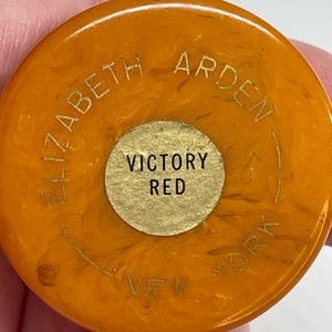 Könnte beinhalten: Ein runder, bernsteinfarbener Kunststoffdeckel mit marmorierter Textur. Der Text ELIZABETH ARDEN NEW YORK ist goldfarben am Rand eingeprägt. Ein kreisförmiger, goldfarbener Aufkleber in der Mitte trägt den schwarzen Aufdruck VICTORY RED.