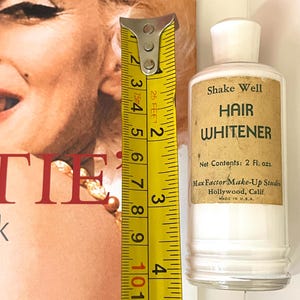 Può includere: Una bottiglia vintage di schiarente per capelli Max Factor con tappo bianco ed etichetta color cuoio con la scritta Shake Well HAIR WHITENER Net Contents: 2 Fl. ozs. Max Factor Make-Up Studio Hollywood, Calif. MADE IN U.S.A. accanto a un metro giallo da 10 cm.