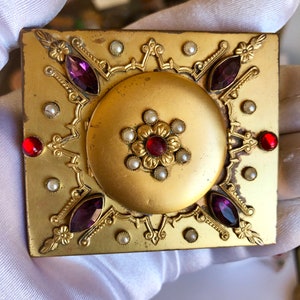 May include: A square gold-tone metal compact case featuring a central circular medallion with a red stone surrounded by small white faux pearls. The corners and edges are decorated with purple marquise-cut stones and red round stones.