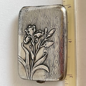 May include: A rectangular silver-toned metal case featuring an embossed iris flower design and a wood grain texture. A ruler placed alongside the object indicates a length of approximately 2.5 inches for the visible portion of the case.