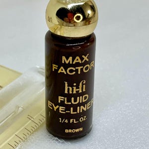 Può includere: Un piccolo flacone marrone di eyeliner liquido Max Factor hi-fi con un tappo dorato lucido. Il testo sull'etichetta recita MAX FACTOR hi-fi FLUID EYE-LINER 1/4 FL. OZ. BROWN. Un righello in plastica trasparente è visibile sul lato sinistro.