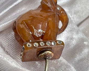 Könnte beinhalten: Eine braune, geschnitzte Elefanten-Anstecknadel mit einem rechteckigen Sockel, der mit einer Reihe kleiner, klarer, funkelnder Strasssteine verziert ist. Die Elefantenfigur sitzt auf dem Sockel, der an einem dünnen, silberfarbenen Metallstift befestigt ist.