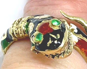 May include: A gold-toned ring shaped like a coiled snake with a textured body. The snake head features black enamel detailing, a central red accent, and two small green stones for eyes. The ring wraps around the finger with a metallic finish.
