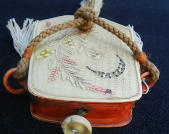 May include: A rectangular off-white case with a textured surface featuring a carved floral motif in pink and yellow, accented by a crescent of small clear stones. The sides are reddish-brown with a flower-shaped knob and a thick braided cord handle.