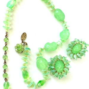 May include: A light green jewelry set consisting of a necklace and a pair of clip-on earrings. The necklace features translucent green beads and small disc-shaped spacers with a metal tag reading Miriam Haskell. The earrings have a central green cabochon surrounded by small green petals.