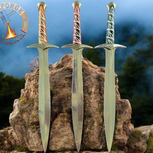 May include: Three decorative swords with ornate handles in orange, pink, and green, resting on a stone. The blades are silver, and the handles feature intricate designs. The Maxforge logo is visible in the upper left corner.