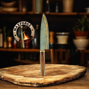 May include: A handcrafted chef's knife with a wooden handle and a patterned blade. The blade has a gold-coloured accent along the edge. The knife is displayed on a wooden cutting board. The logo "MAXFORGE" is visible in the background.