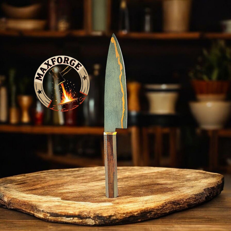 May include: A handcrafted chef's knife with a wooden handle and a patterned blade. The blade has a gold-coloured accent along the edge. The knife is displayed on a wooden cutting board. The logo "MAXFORGE" is visible in the background.