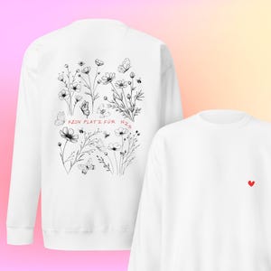 Op de afbeelding: Twee witte sweaters met ronde hals. De linker sweater toont een zwarte lijntekening van wilde bloemen, vlinders en bijen met de rode tekst "KEIN PLATZ FÜR NZS". De rechter sweater is voorzien van een klein, rood hartje in het midden.