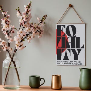 Könnte beinhalten: Ein Stillleben mit einem gerahmten Druck mit dem Wort "EQUALITY" in fetten schwarzen und roten Buchstaben, mit dem Text "STARTS AT THE KITCHEN TABLE." Eine klare Glasvase hält blühende Zweige. Eine grüne Tasse, ein goldfarbener Becher und ein grüner Krug stehen auf einer Holzoberfläche.