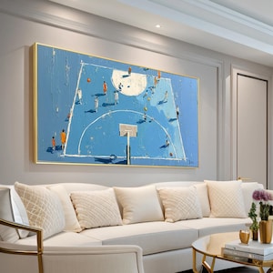 May include: A large, rectangular painting depicts a basketball court in shades of blue with players in action. The artwork features a textured, impasto style with a gold frame, creating a modern aesthetic. The painting hangs above a white sofa in a living room setting.