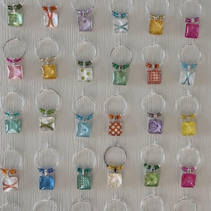 May include: A collection of colorful wine charms with square pendants and beaded accents. Each charm features a different design, including polka dots, swirls, and geometric patterns, in shades of pink, green, orange, and blue. The charms are attached to silver-toned hoops.