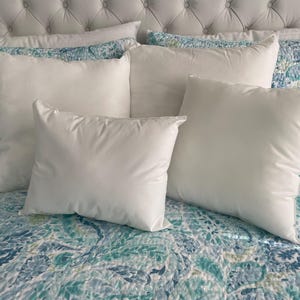 May include: A collection of white square pillows of varying sizes arranged on a bed. The pillows are fluffy and have a smooth texture. The bedspread features a blue and teal floral pattern, and the headboard is a light gray, button-tufted design.