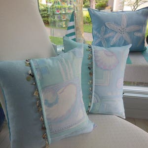 May include: Three decorative pillows with a light blue background. The pillows have different designs, including a seashell pattern, a starfish pattern, and a more abstract pattern with a light blue and white color scheme. The pillows are on a white chair with a light blue cushion.