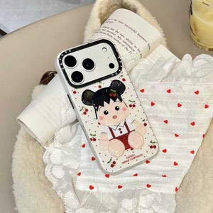 May include: White iPhone case with a cartoon girl design. The case features a black camera module and a pattern of cherries. The girl is wearing a red outfit. The text "CAFADU 100% OF COMPANION" is printed on the case.