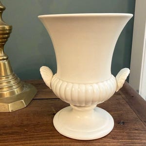 May include: A cream-coloured ceramic urn-style vase featuring a wide, flared rim, a ribbed body, and two decorative handles shaped like seashells. The vessel stands on a circular pedestal base and has a smooth, matte finish throughout its structure.